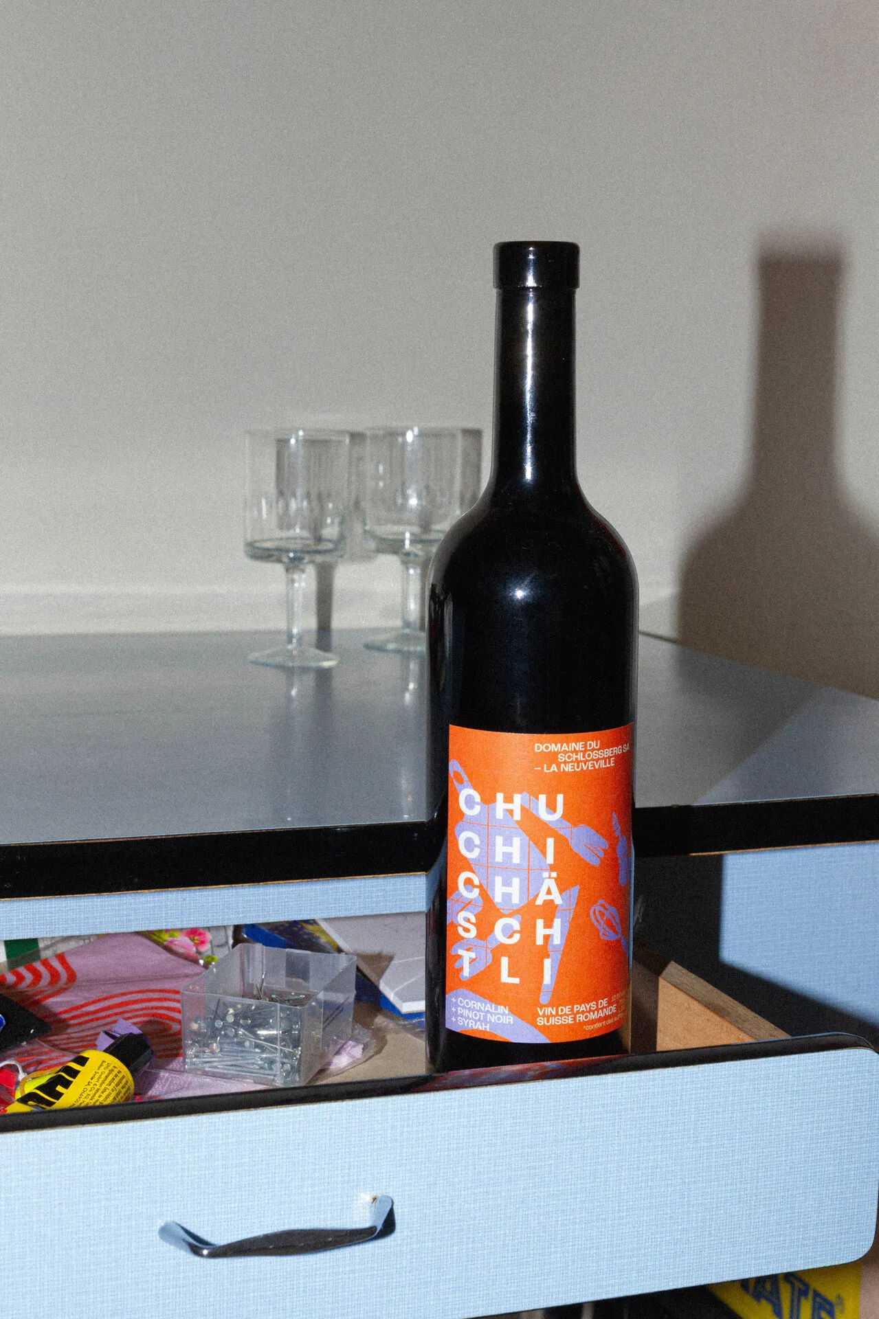 Bouteille de vin à l’étiquette orange CHUCHICHASCHTLI posée devant un tiroir de cuisine ouvert